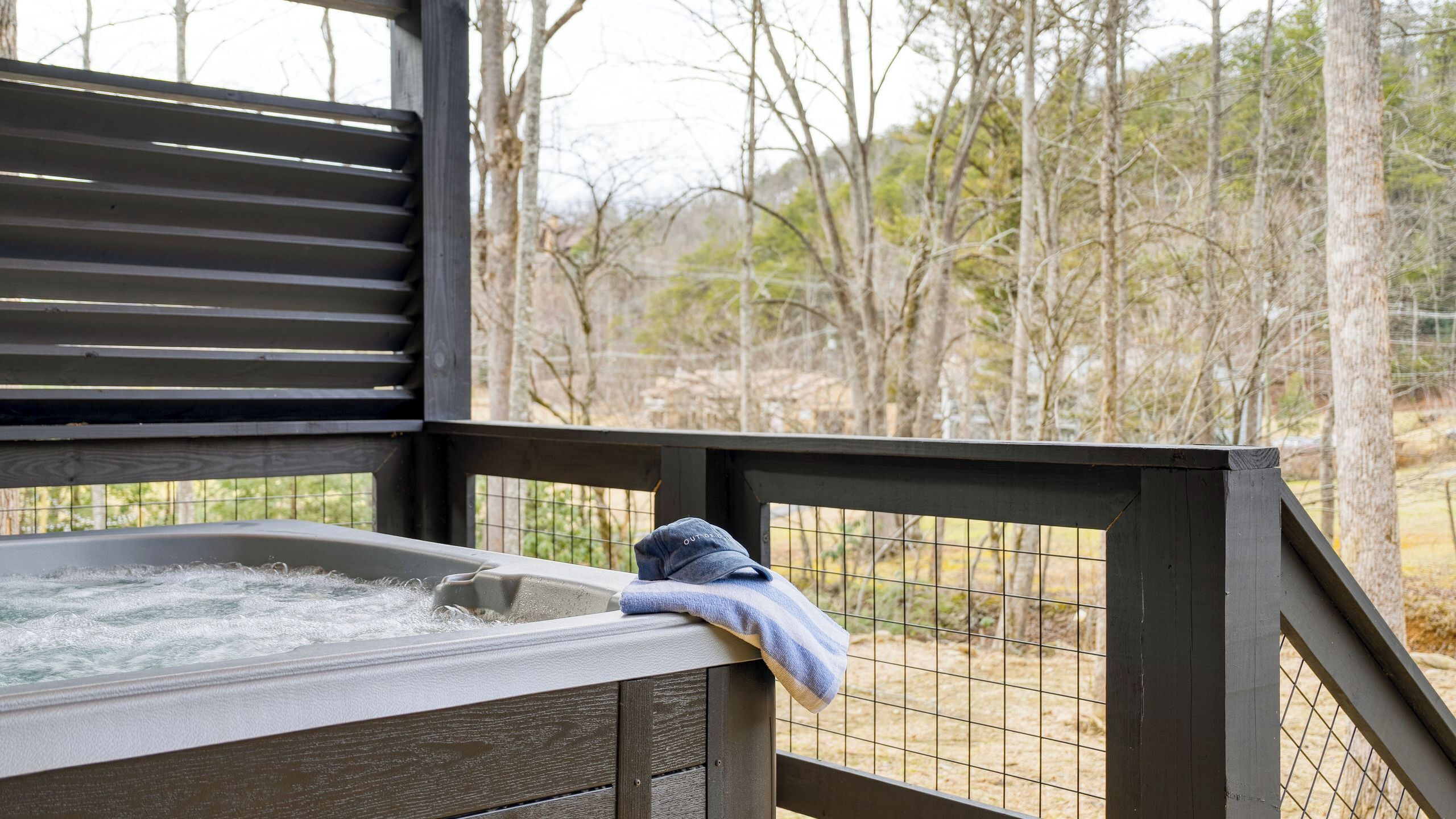 A hot tub with a towel on the edge, surrounded by a wooden deck and a wire fence, overlooking a wooded area.