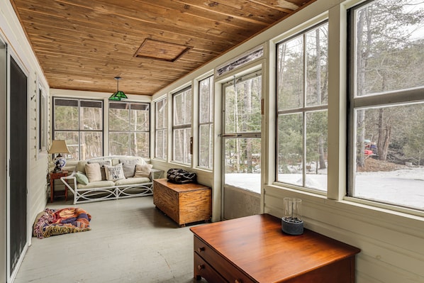 Una habitación con techo de madera, ventanas grandes, un sofá blanco, un cofre de madera y una mesita con una lámpara.