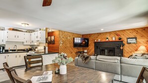 A modern kitchen with white cabinets, a dining table with chairs, a grey sofa, a fireplace, and a mounted TV. - Riverfront Cabin w/ Hot Tub in Schroon Lake! (Schroon Lake)
