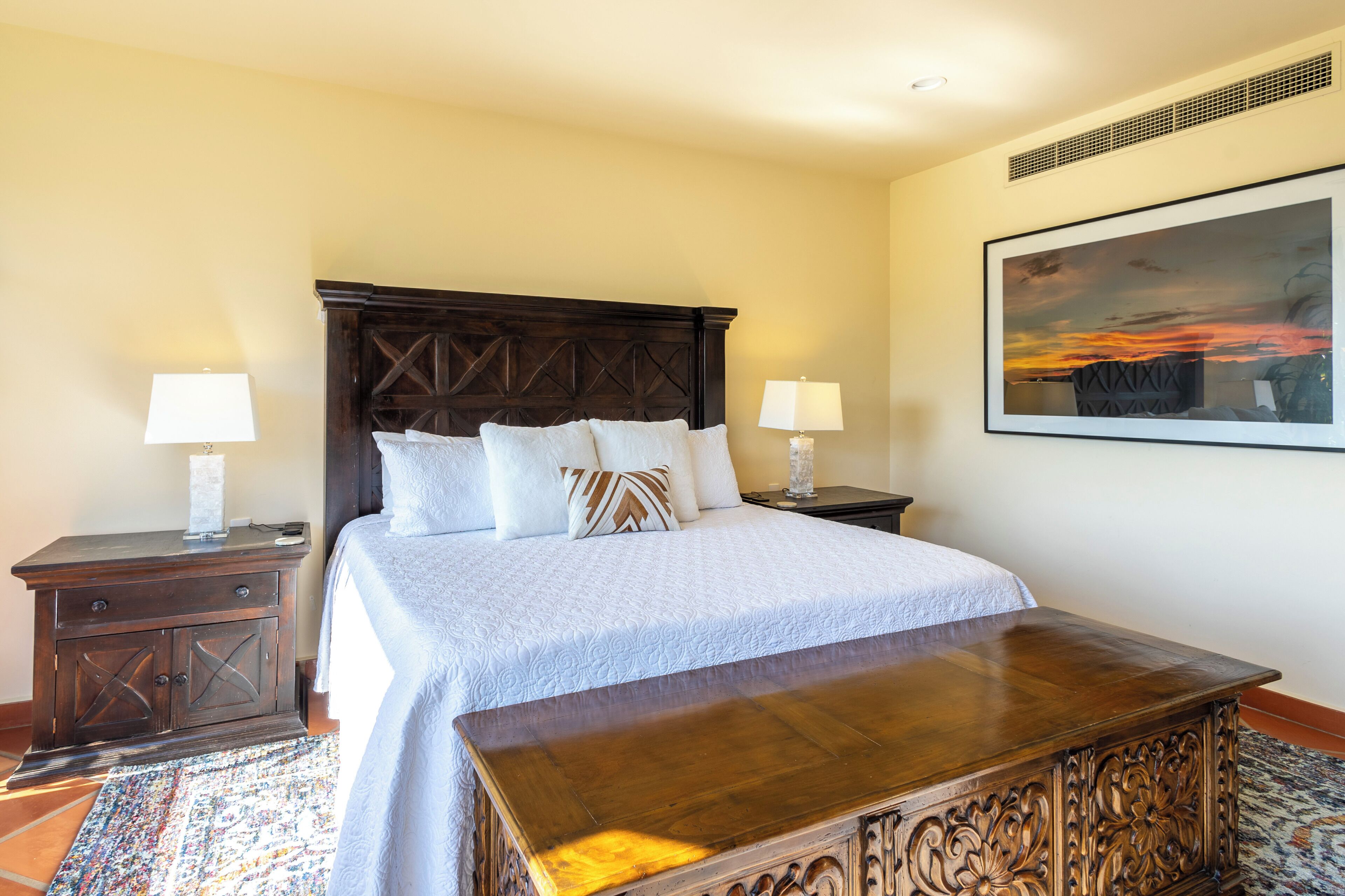 A bedroom with a large bed, two bedside tables, a wooden bench, a patterned rug, and a framed picture on the wall.