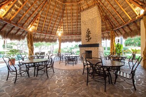 An outdoor patio with stone flooring, metal tables, and chairs, surrounded by a thatched roof structure.