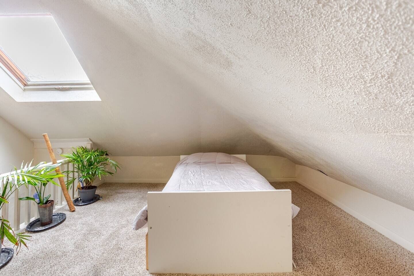 Een kleine, witte kamer met een enkel bed, schuine wanden, een dakraam en verschillende potplanten.