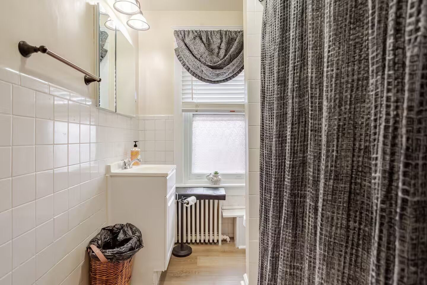 A bathroom with white tiled walls, a sink with a soap dispenser, a radiator, and a window with a patterned curtain.