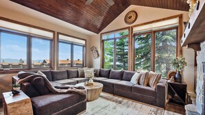 A cozy living room with a sectional sofa, a wooden coffee table, and large windows offering a view of trees and mountains. - Snow Therapy Chalet Ski In Out & Hot Tub (Big White)