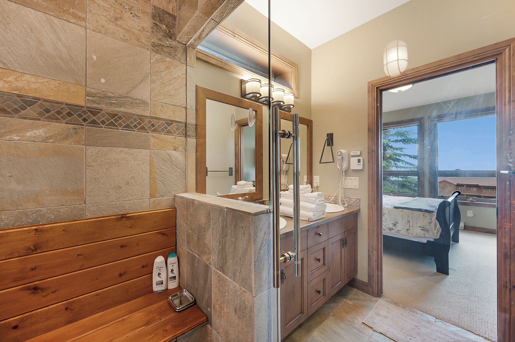 A bathroom with stone tiles, wooden benches, a vanity with a mirror, and a view of a balcony.
