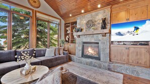 A cozy living room with a stone fireplace, large windows, and a television displaying a snowy mountain scene. - Snow Therapy Chalet Ski In Out & Hot Tub (Big White)