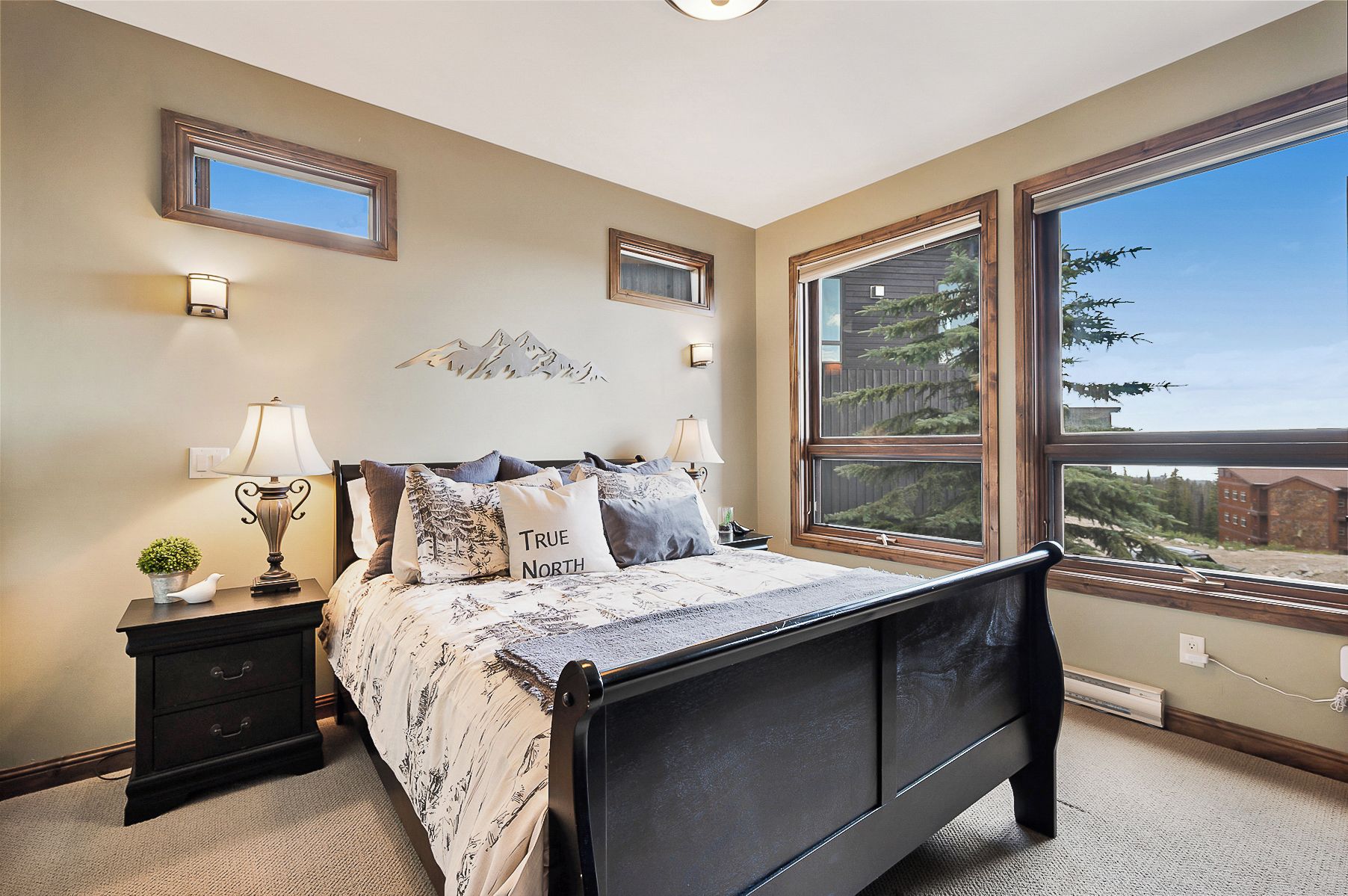 Une chambre à coucher comprenant un lit, des tables de chevet, des lampes, un miroir fixé au mur et une grande fenêtre donnant sur des arbres et un immeuble.
