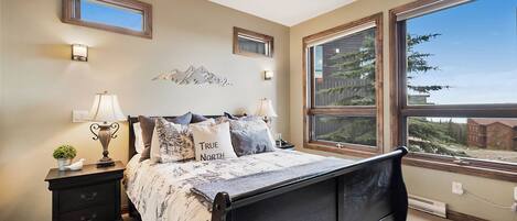 Une chambre à coucher comprenant un lit, des tables de chevet, des lampes, un miroir fixé au mur et une grande fenêtre donnant sur des arbres et un immeuble.