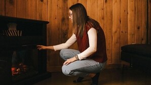 A person crouching near a fireplace in a room with wooden walls and a horse painting. - Domaine Yamaska (Bromont)