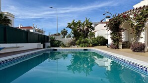 Una piscina con borde de azulejos, rodeada de vegetación y un edificio blanco con techo de tejas rojas.
