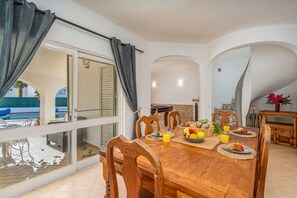Un espace repas avec une table en bois, des chaises et une vue sur une piscine et un coin salon extérieur.
