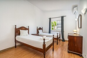 Une chambre d’hôtel avec deux lits en bois, une petite table de chevet, une commode, une coiffeuse en bois, une fenêtre avec des rideaux et un climatiseur fixé au mur.