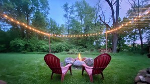 Two Adirondack chairs facing a fire pit surrounded by a string of lights, set in a grassy outdoor area with trees in the background.
