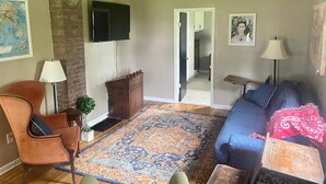 A living room with a blue sofa, a brown armchair, a wooden cabinet, a TV mounted on the wall, and a patterned rug.