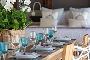 A dining table set with blue glassware and white plates, a wicker basket, and white chairs in a room with a bed and pillows in the background. - Chic Baan Capo - Luxury Four Bedroom Beachfront Villa (Koh Samui)