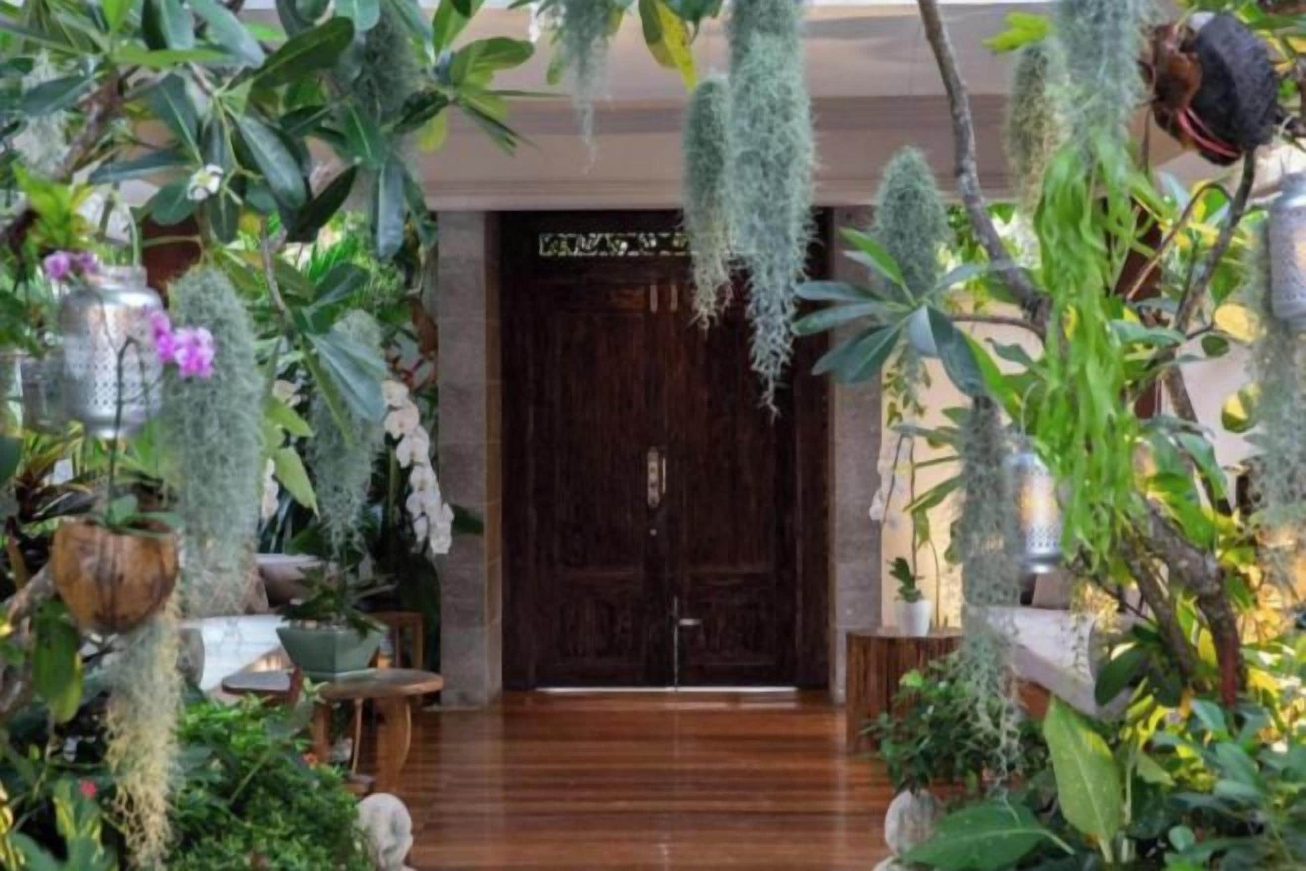 A hotel lobby with a wooden floor, a dark wooden door, and various plants and greenery.