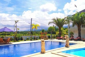 A swimming pool with two stone pillars, surrounded by lounge chairs and umbrellas, with a view of a mountain and palm trees in the background. - Puri Lovina Hotel (Bali)
