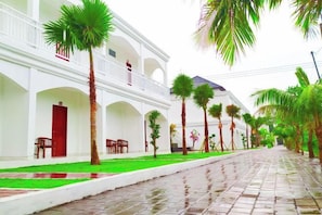 A white building with a covered walkway, palm trees, and a wet pavement. - Puri Lovina Hotel (Bali)