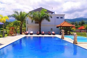 A swimming pool with lounge chairs, a palm tree, and a building in the background. - Puri Lovina Hotel (Bali)