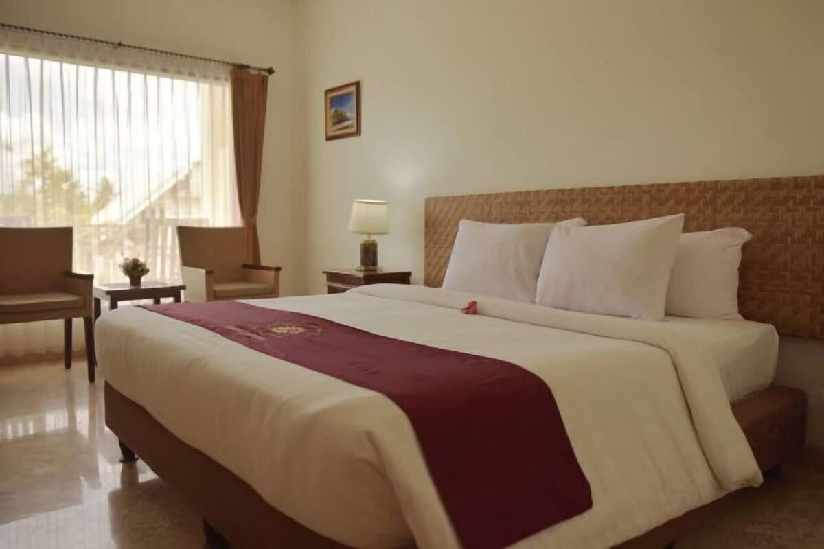 Ein Hotelzimmer mit einem großen Bett, einem Nachttisch mit Lampe, einem Sessel und einem Fenster mit Vorhängen.