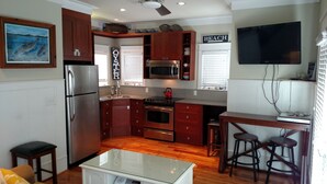 Cocina compacta con armarios de madera, un refrigerador de acero inoxidable, un microondas y una estufa. Cuenta con una barra con taburetes y un televisor montado en la pared.