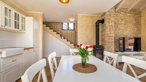 A dining area with a white table, white chairs, and a vase of flowers. There is a staircase leading to another level, a brick wall, and a television on a stand. - Semi-detached holiday house DIVNA with private pool (Pinezići)