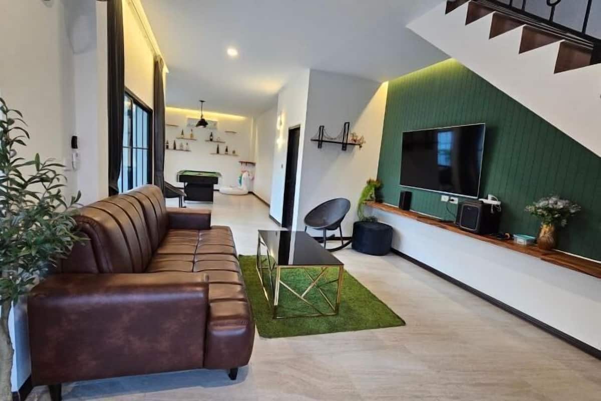 A modern living room with a brown leather sofa, a glass coffee table, a TV mounted on a green wall, and a staircase.