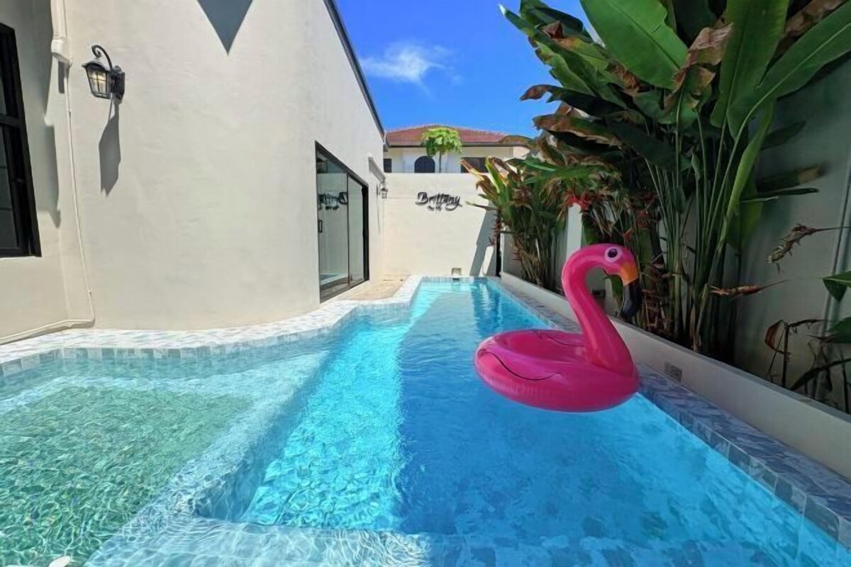 A swimming pool with a pink flamingo float, surrounded by plants and a building with a white exterior.