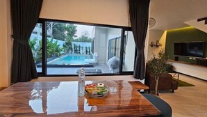 Un salon moderne doté d’une grande porte vitrée donnant sur une piscine, d’une table à manger avec un saladier de fruits et d’un téléviseur fixé sur un mur vert.