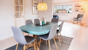 A dining area with a round table, grey chairs, a woven pendant light, and a view of the kitchen through an open door. - Villa Holstebro (Holstebro)