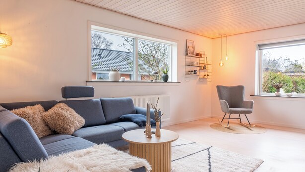 A modern living room with a grey sofa, a round coffee table, and a white furry ottoman. There is a window with a view of a house and trees, and a grey chair with a wooden base. - Villa Holstebro (Holstebro)