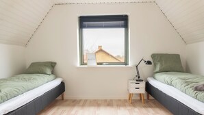 A small, well-lit room with two beds, a nightstand, and a window with a view of a house. - Villa Holstebro (Holstebro)