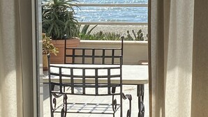 Blick durch ein Fenster auf einen Strand und das Meer, mit einem Metallstuhl und Tisch im Vordergrund.