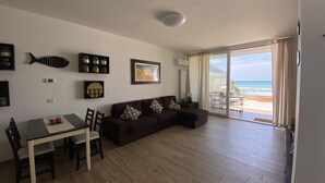 Ein Wohnzimmer mit Holzboden, einem dunkelbraunen Sofa, einem Esstisch mit Stühlen und Blick auf den Strand durch große Glasfenster.