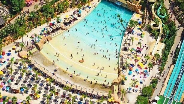 Eine Luftaufnahme eines Resorts mit großem Pool, mehreren Wasserrutschen und einem Strandbereich mit Liegestühlen und Sonnenschirmen.