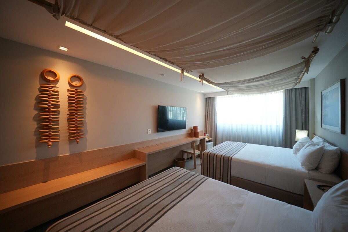 A hotel room with two beds, a flat-screen TV, a wooden bench, and a large window with curtains.