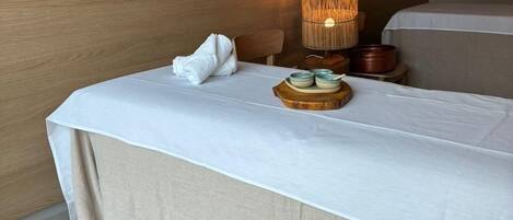 Una habitación de hotel con una cama, un taburete de madera, una mesa con una bandeja de objetos y una lámpara.