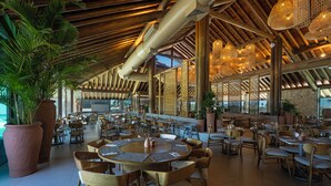 A spacious indoor dining area with round tables and chairs, large potted plants, and a high wooden ceiling with exposed beams and hanging lights.
