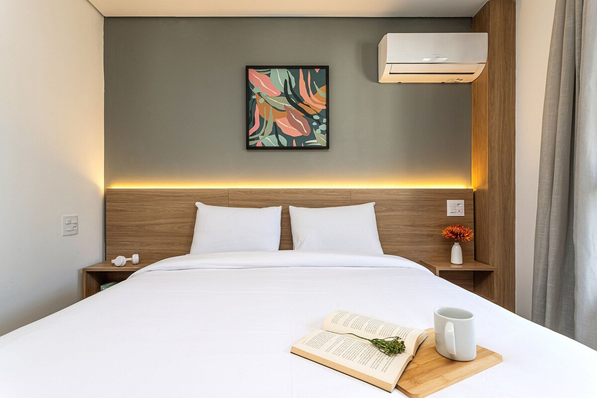 A neatly made bed with white linens, a bedside table with a book and mug, a wall-mounted air conditioner, and a painting above the bed.