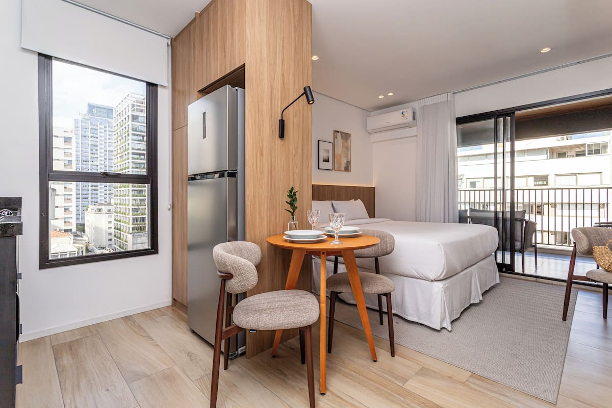 Une chambre d’hôtel moderne équipée d’un lit, d’une table à manger avec des chaises, d’un réfrigérateur et offrant une vue sur le paysage urbain grâce à de grandes fenêtres.