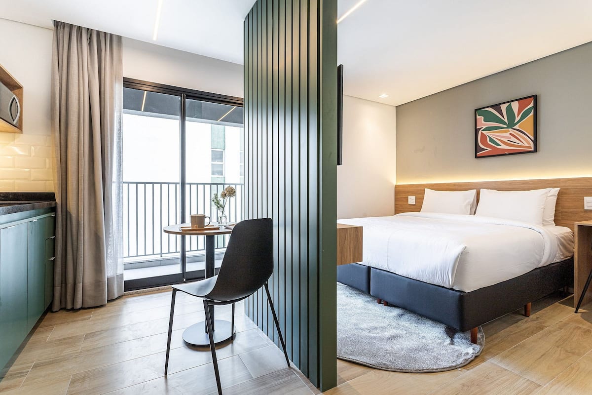 Une chambre d’hôtel avec un lit, une chaise, une petite table, une fenêtre avec des rideaux et un balcon.
