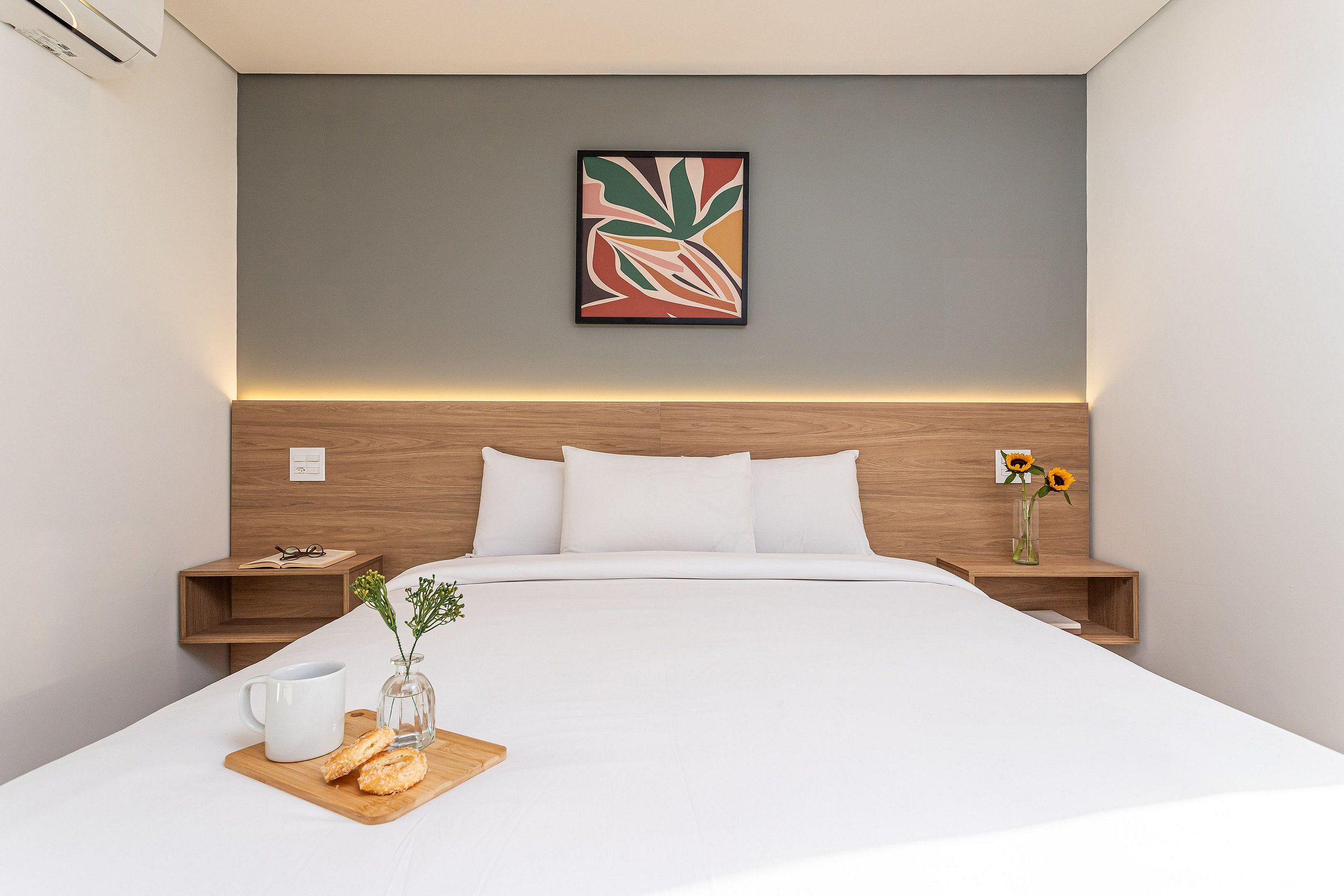 A modern hotel room with a neatly made bed, a bedside table with a vase of sunflowers, and a framed abstract painting on the wall.