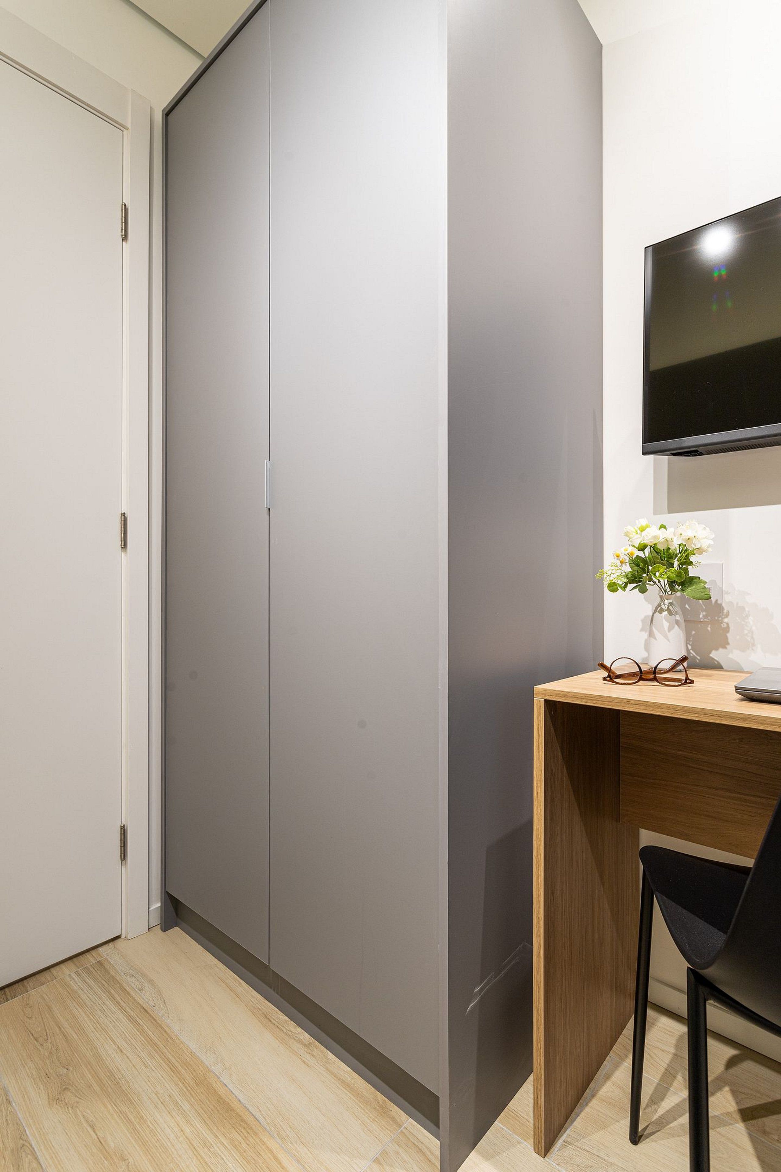 Une chambre comprenant une armoire, un bureau agrémenté d’un vase de fleurs, une chaise et un téléviseur à écran plat fixé au mur.