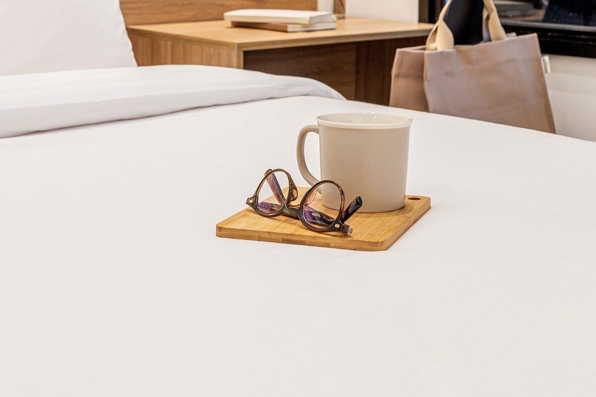 A hotel room with a bed, a wooden nightstand, a white pillow, a white mug, and a pair of sunglasses on a wooden coaster.