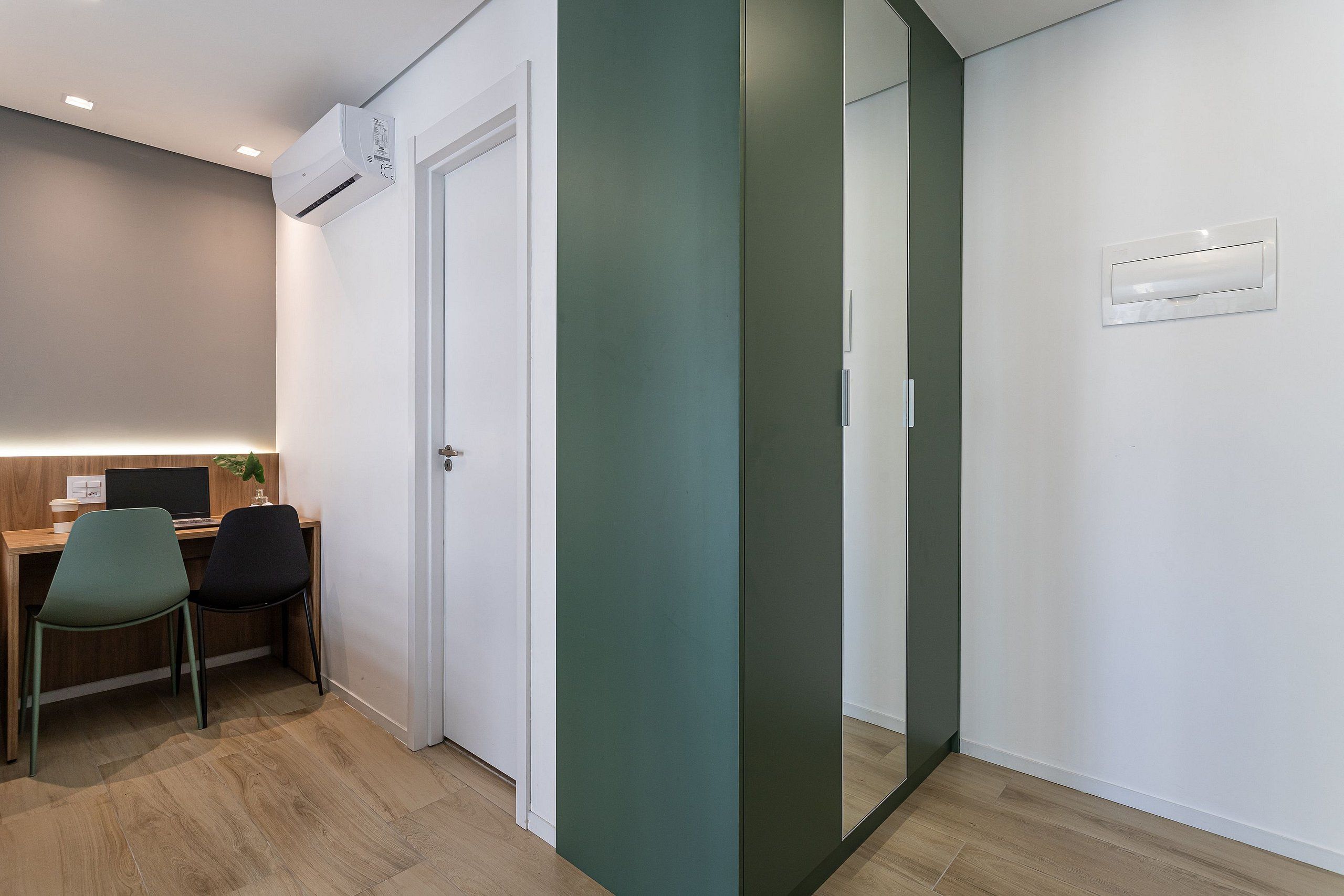 Ein Zimmer mit Schreibtisch, zwei Stühlen, einer Klimaanlage, einer Tür und einem wandmontierten Lichtschalter.