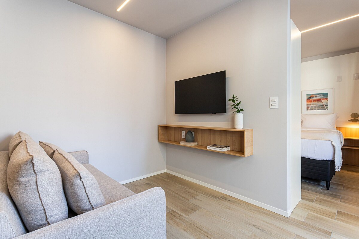 A modern living room with a flat-screen TV mounted on the wall, a wooden shelf with decorative items, a grey sofa with cushions, and a wooden floor.