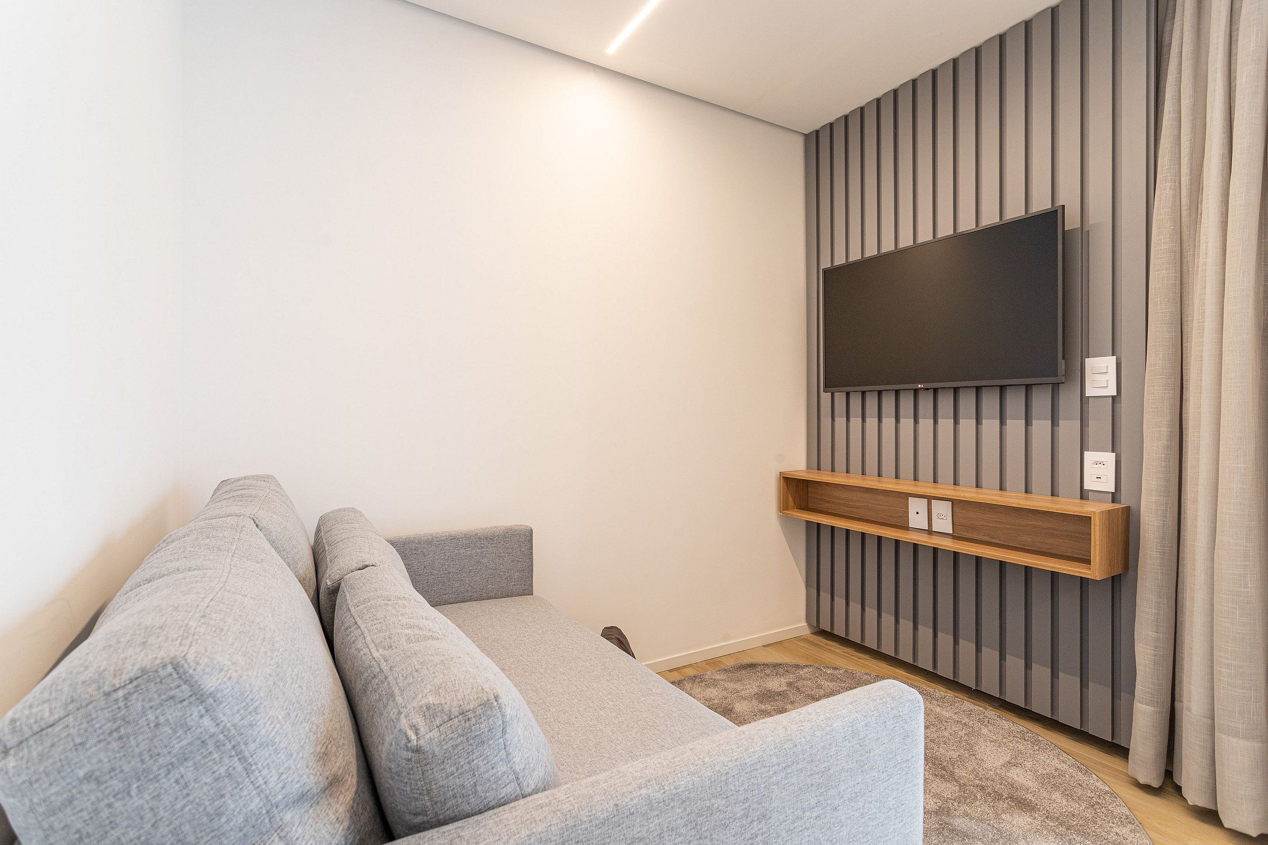 A modern living room with a grey sofa, a mounted TV, and a wooden shelf.