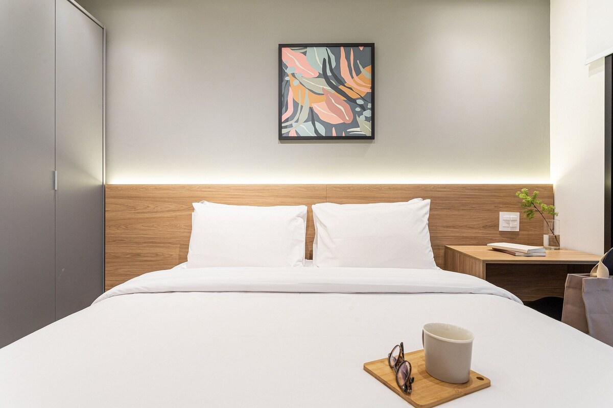 A hotel room with a bed, a nightstand, a framed abstract painting, and a bedside table with a plant.