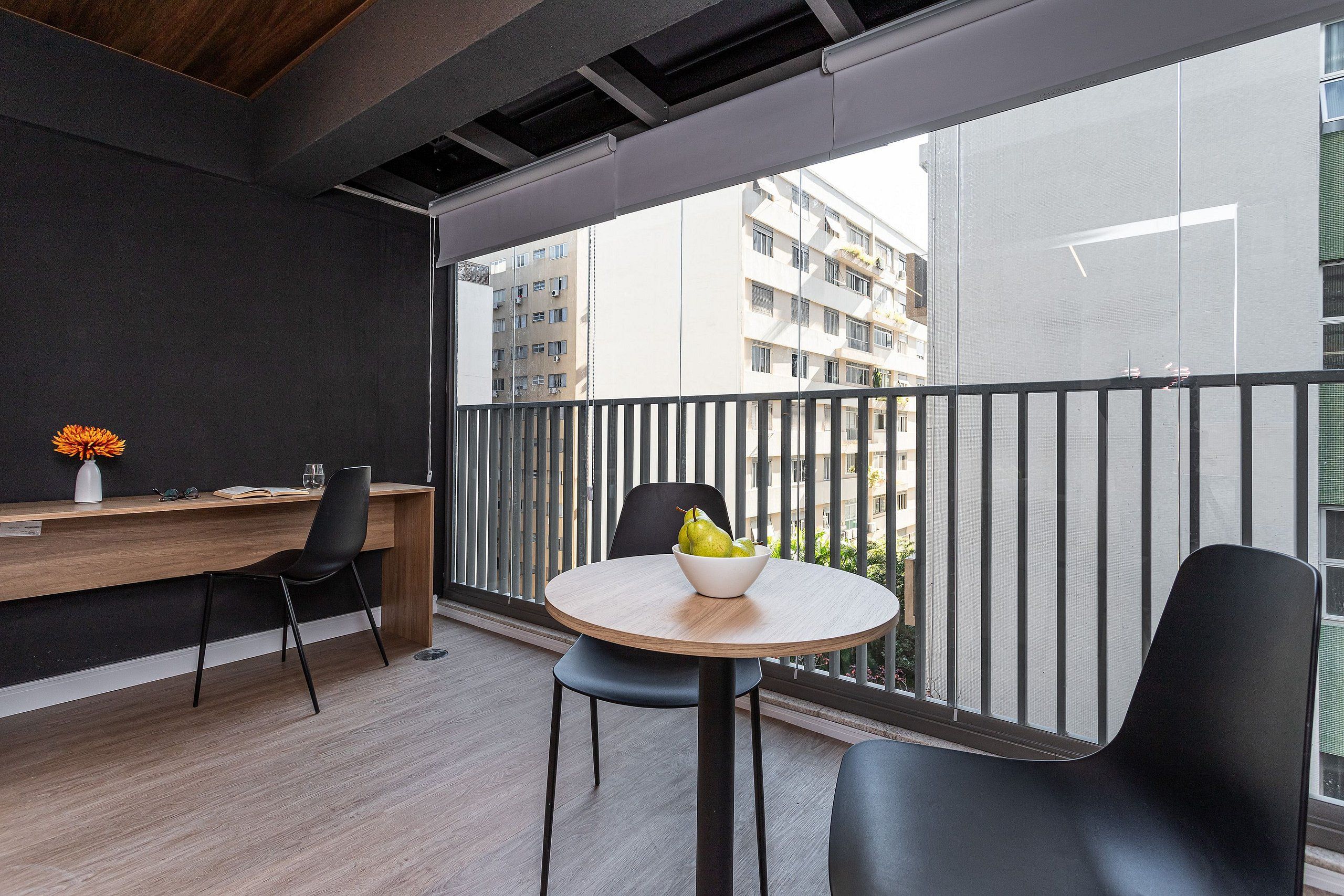 Un balcón moderno con un escritorio de madera, una mesa redonda con un frutero y dos sillas negras.