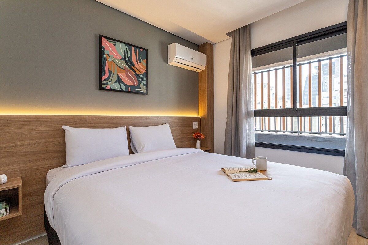 A hotel room with a bed, a bedside table, a window with curtains, a wall-mounted air conditioner, and a painting.